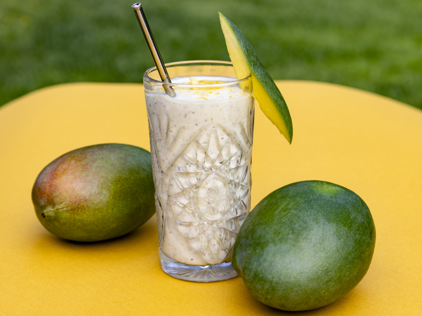 Nápoj Mango lassi s chia semínky servírovaný v průhledné skleničce s lesklým brčkem, která je ozdobená kouskem manga. Po obou stranách skleničky jsou manga.