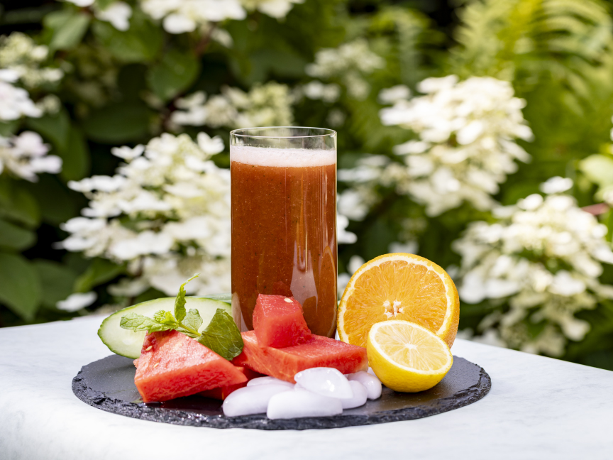 Smoothie z červeného melounu ve skleničce, v její bezprostřední blízkosti jsou kousky melounu a pomeranče. Sklenička stojí venku na stole na bílém ubruse.